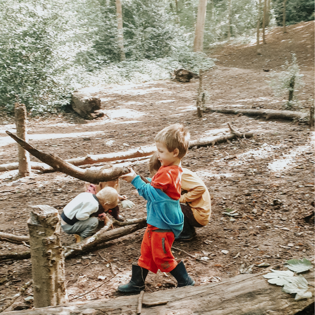 Children play in the forest. One boy in red trousers and a blue jacket carries a large stick