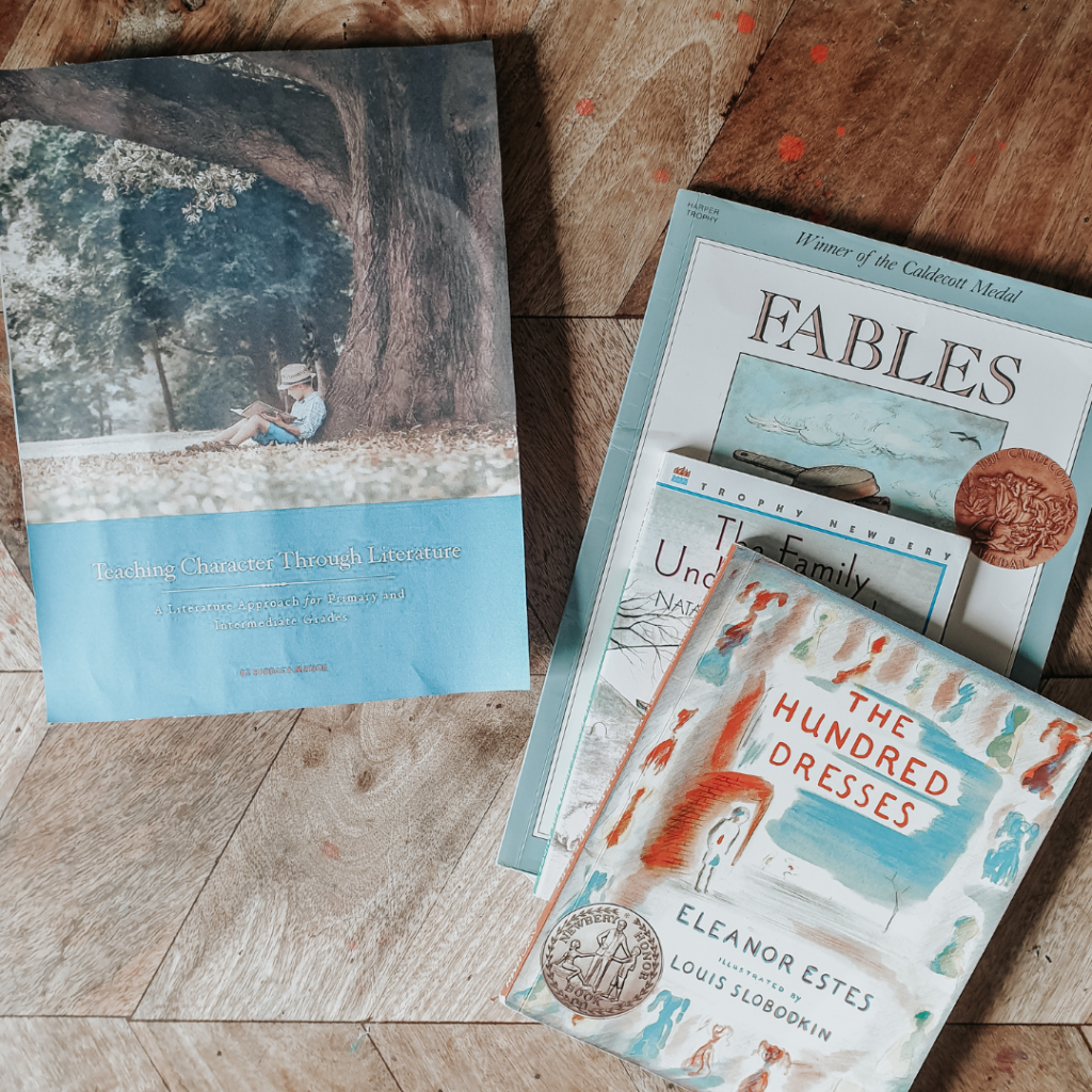 A collection of books on a table. The book on the top is called The Hundred Dresses, and the curriculum next to the books is titled Teaching Character Through Literature