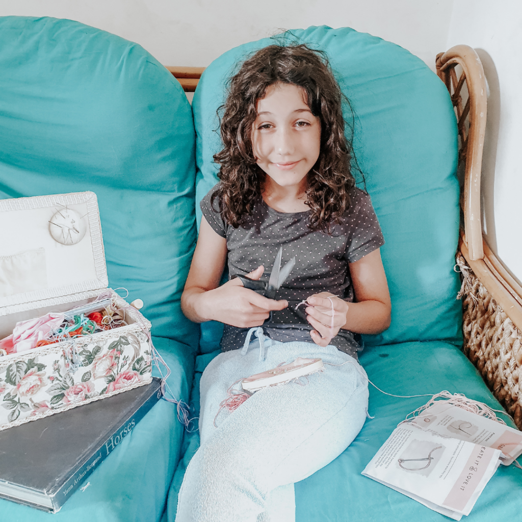 Girl sits on a sofa whilst sewing with an embroidery kit next to her